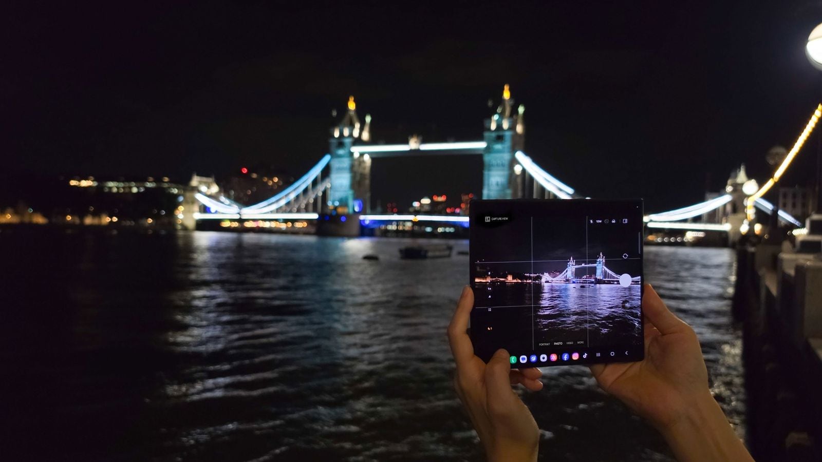 Ruky držiace rozložený Samsung Galaxy Z Fold s otvorenou aplikáciou fotoaparátu, v pozadí nočný Tower Bridge